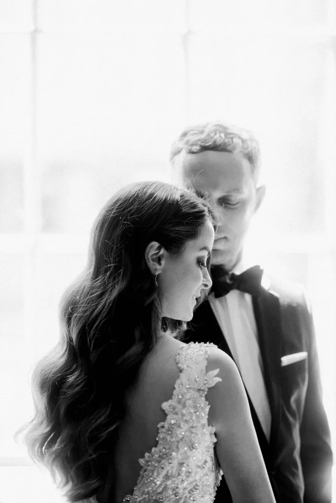 A monochrome image of a bride and groom in elegant wedding attire. the bride, whose cascading hair is detailed, stands in the foreground, while the groom gazes at her from behind. captured by Seattle Wedding Photographers Jenn Tai & Co