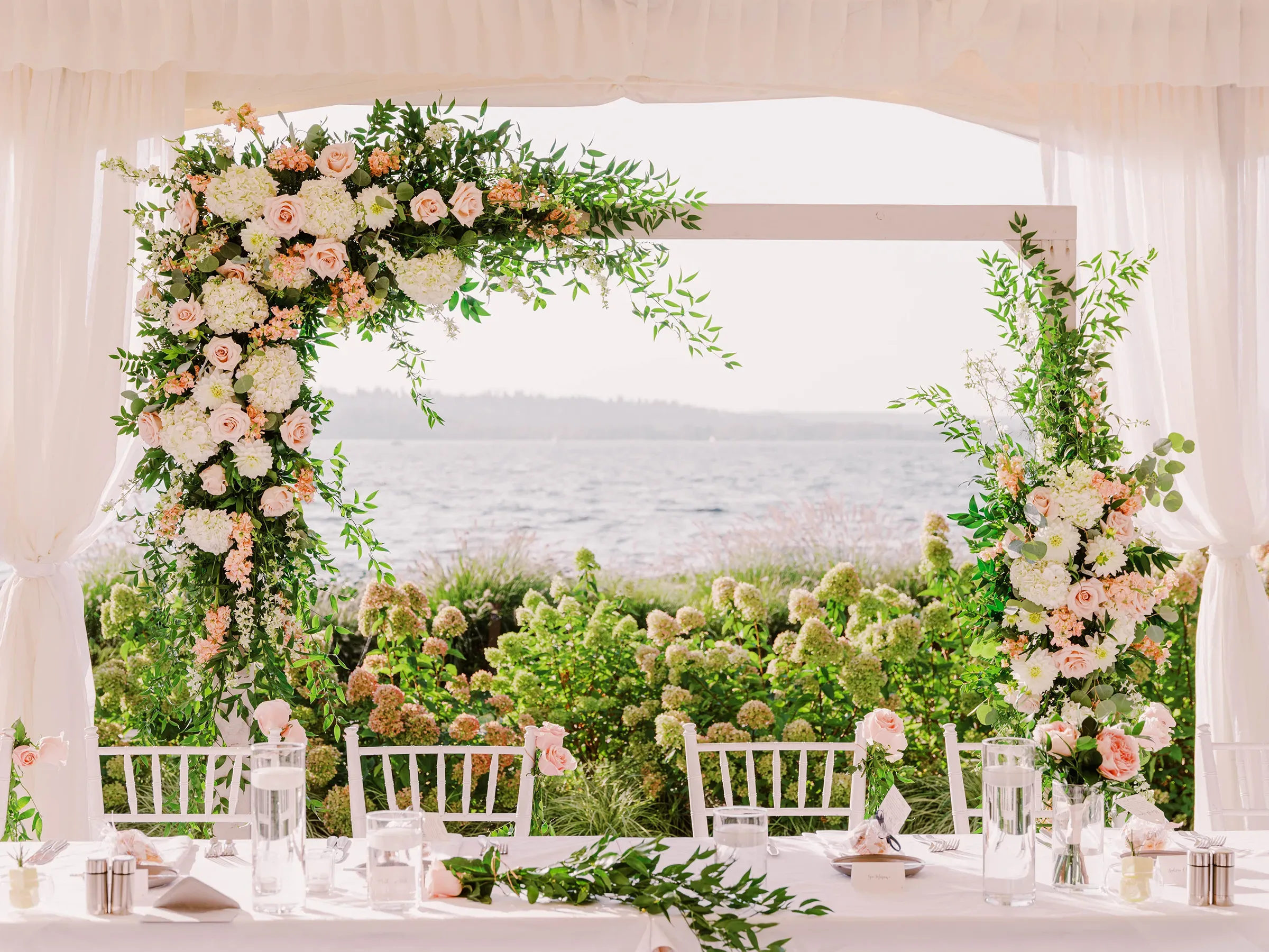 An elegant outdoor wedding setup by the sea featuring a floral arch, white draped chairs, and meticulously arranged dining tables adorned with pink and white flowers. captured by Seattle Wedding Photographers Jenn Tai & Co