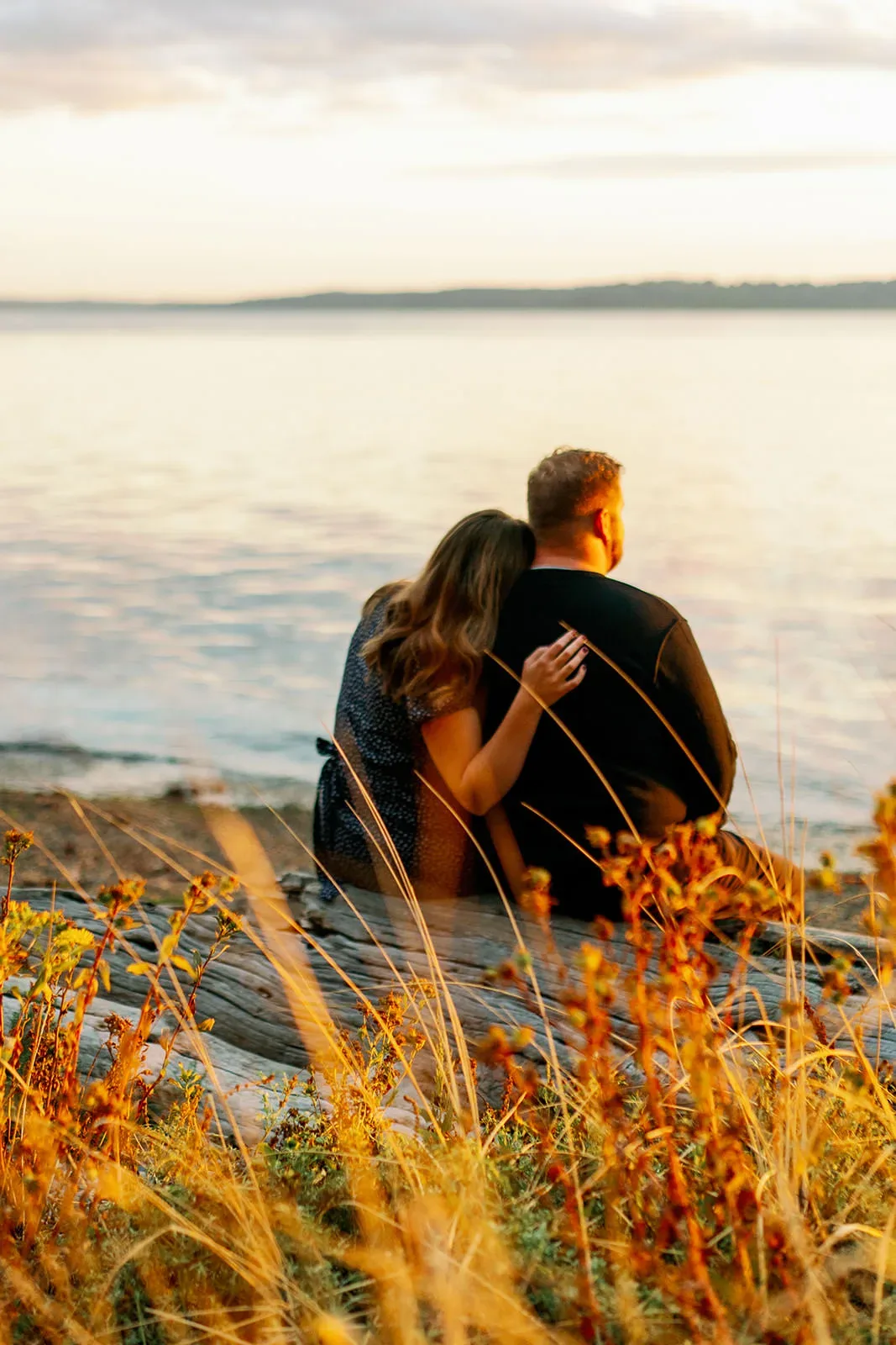 Seattle engagement photos in Lincoln Park A couple sits on a log by the water's edge, with the woman resting her head on the man's shoulder. The scene is bathed in the warm, golden light of sunset. Tall grasses and wildflowers frame the foreground, adding to the serene atmosphere. captured by Seattle Wedding Photographers Jenn Tai & Co